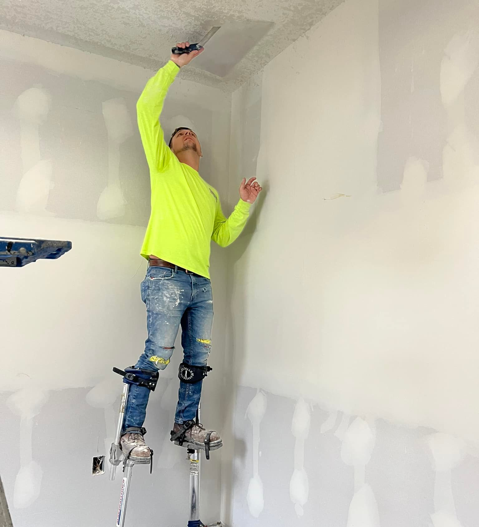 Man on stilts, wearing neon shirt, using a trowel to apply drywall compound to a wall and ceiling.