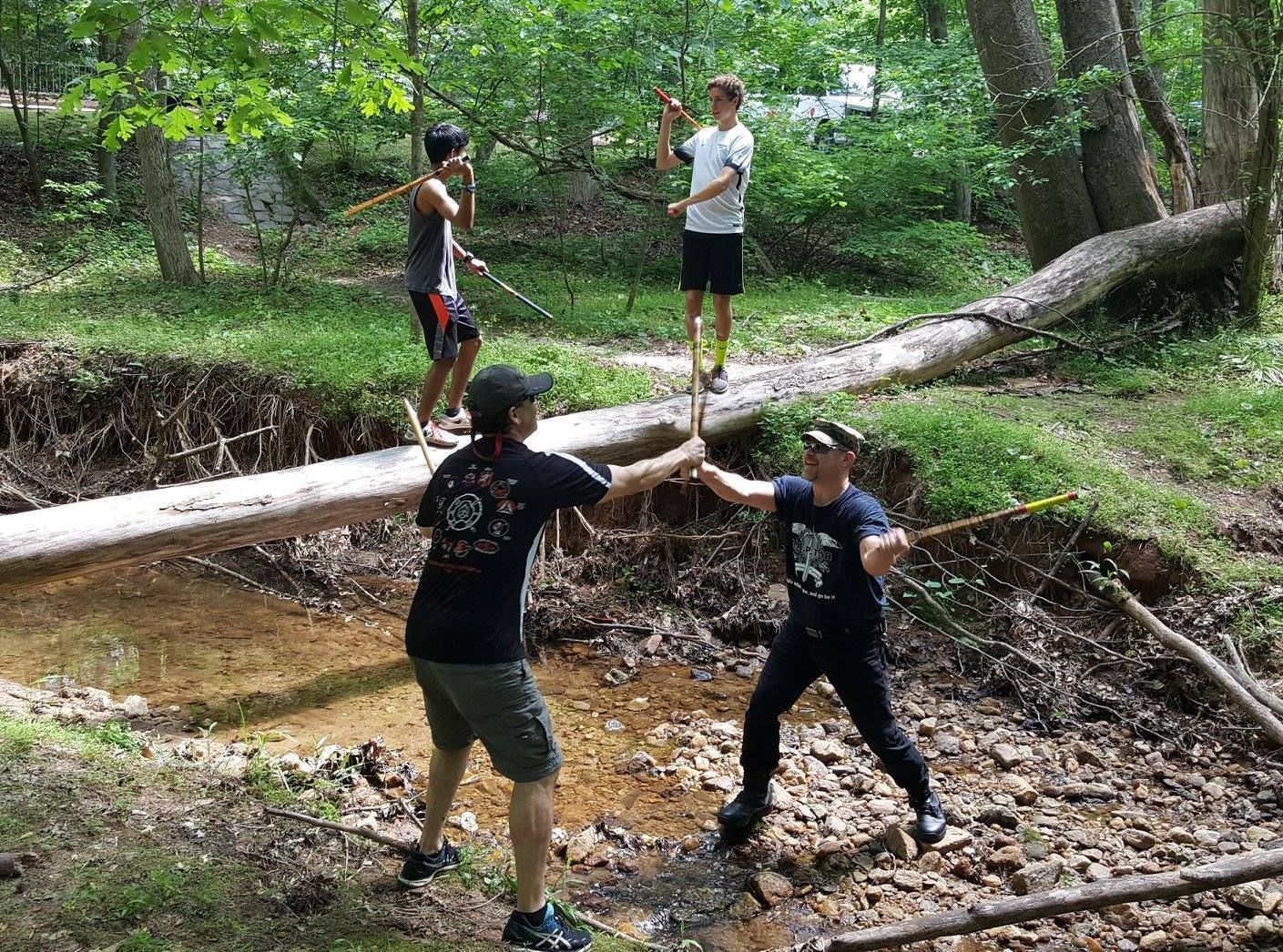 a group of people are standing on a log over a stream in the woods .