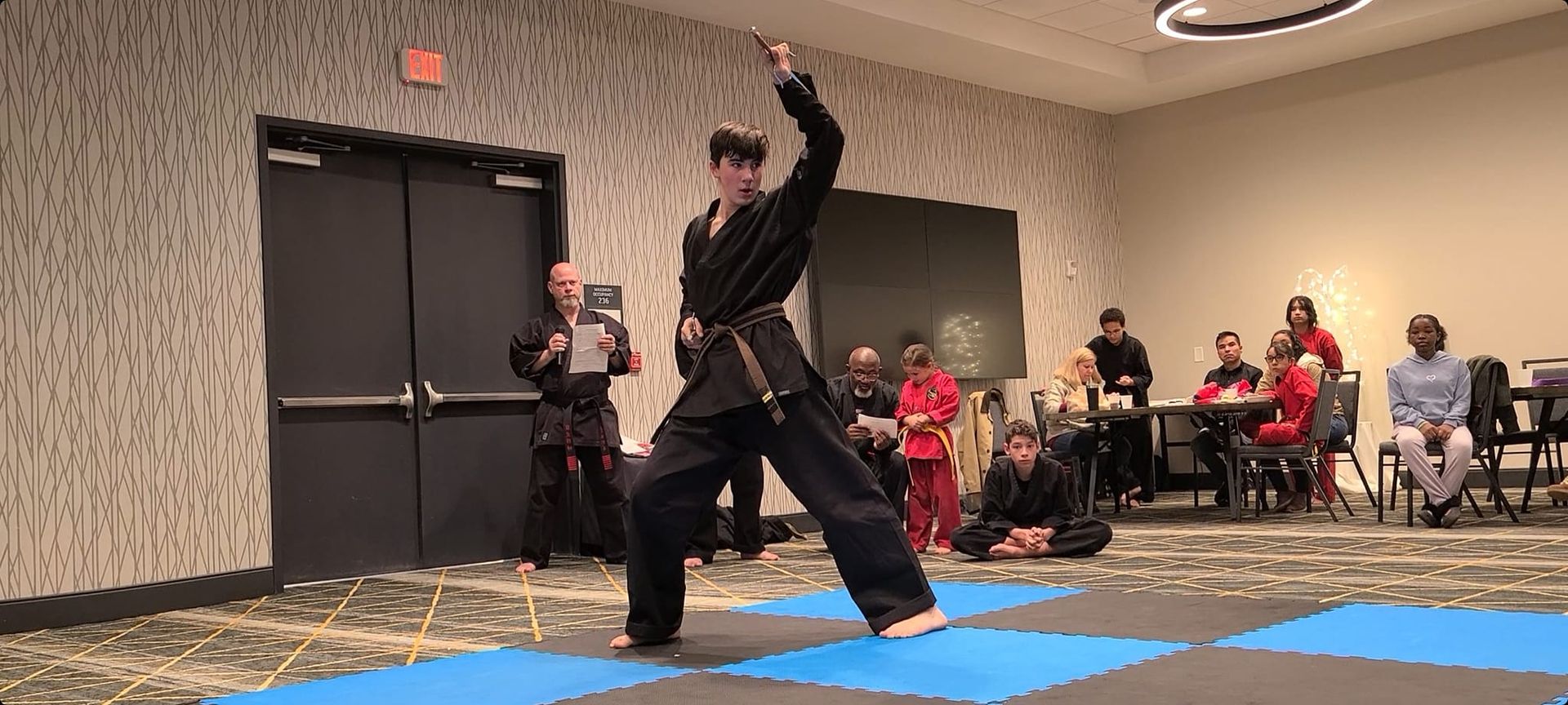 a man in a black karate uniform is standing on a blue mat in a room .