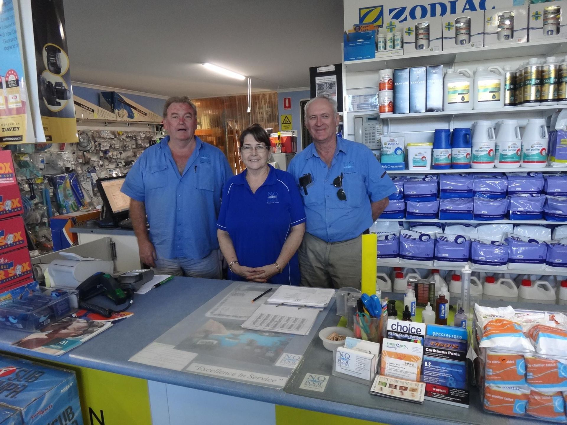 Three people behind a counter in a store, likely a pool supply shop. Blue shirts, supplies on shelves, and smiling.— NQ Pool Supplies in Smithfield, QLD