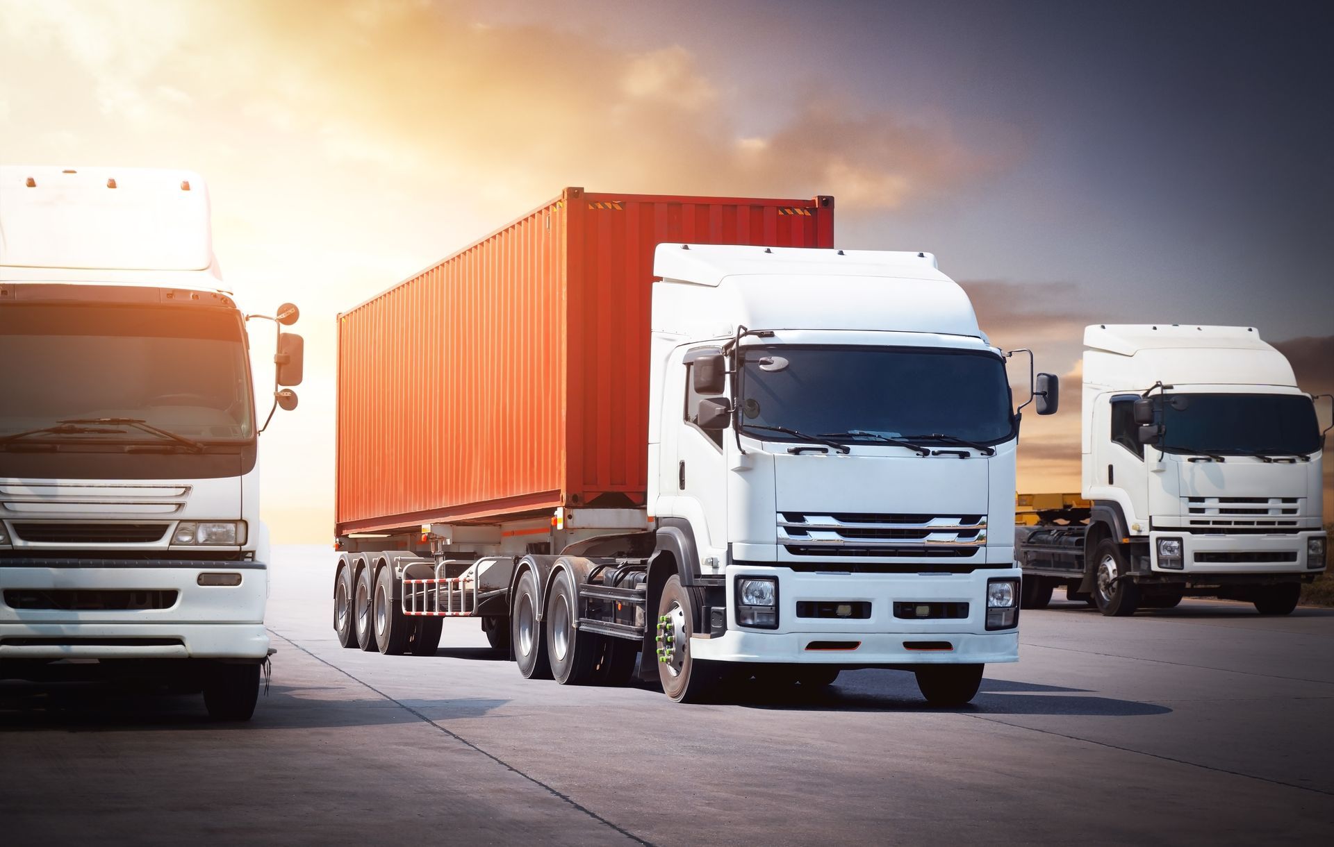 Three white semi-trucks on a paved road, one towing a red cargo container, against a sunset sky.