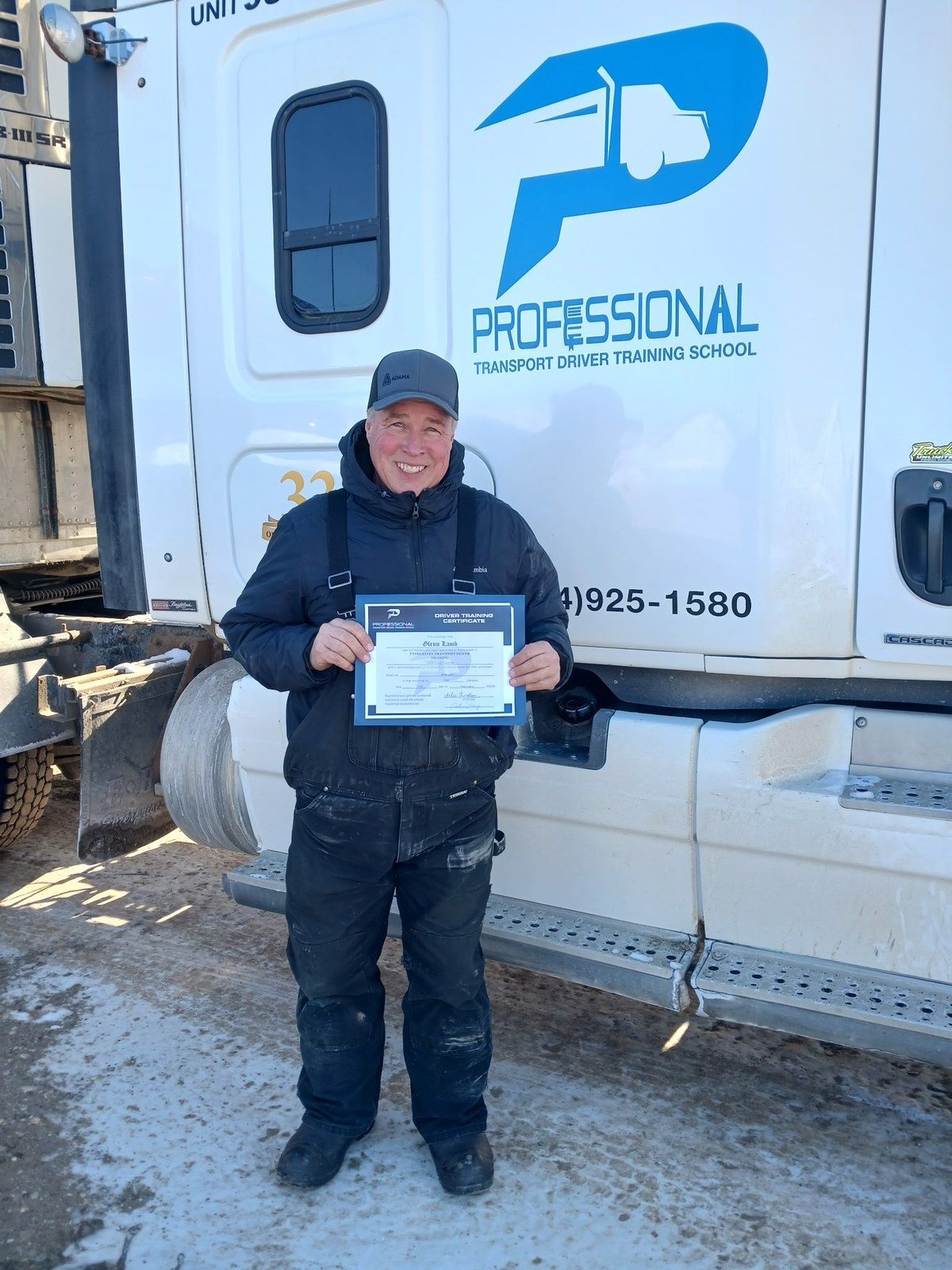 A smiling person in work clothes stands in front of a white semi-truck, holding an award certificate.