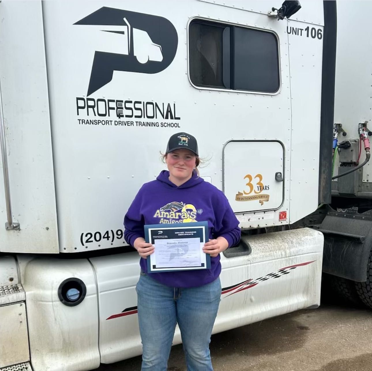 A woman in a purple shirt is standing in front of a semi truck holding a certificate.