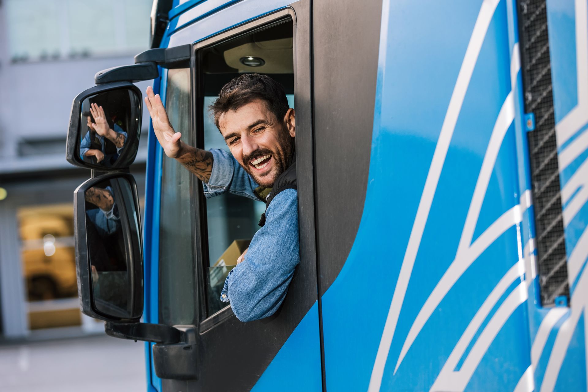 Truck driver waves enthusiastically from the window of a blue and white truck.