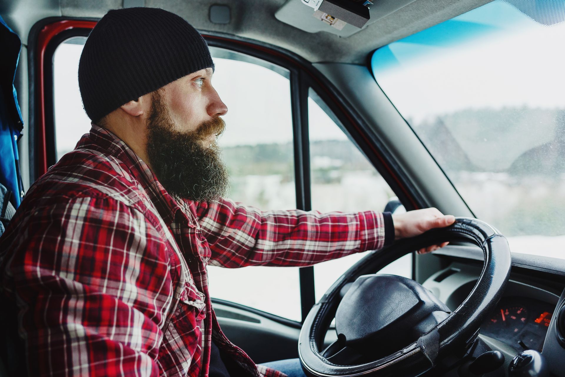 Man with a beard wearing a plaid shirt and beanie, driving a vehicle on a snowy day.