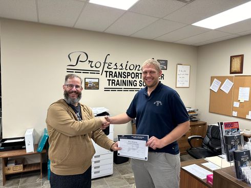 Two men are shaking hands in a room with a sign on the wall that says professor transport training center.