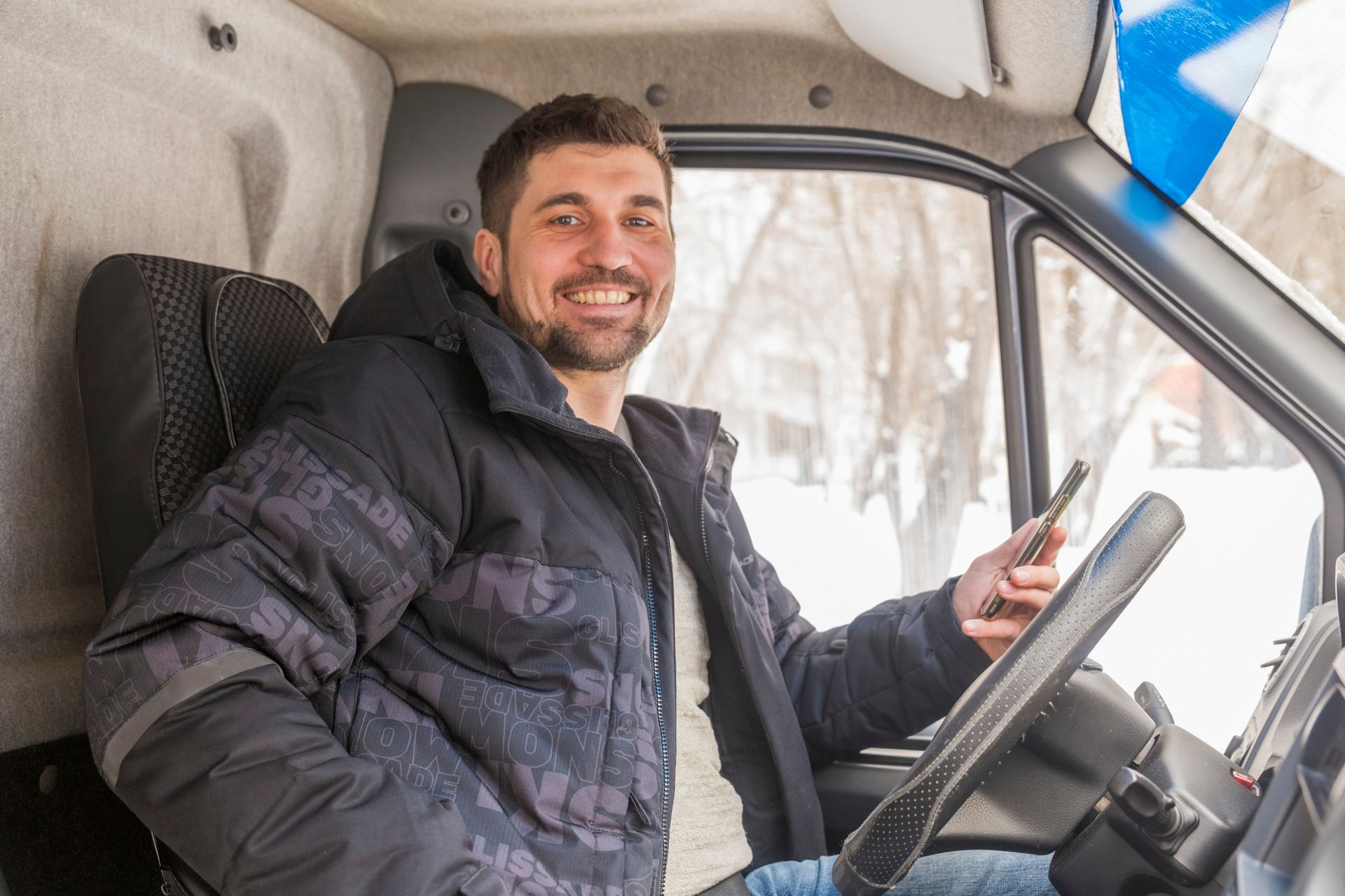 Man in a winter coat smiles while sitting in a truck and holding a cell phone.