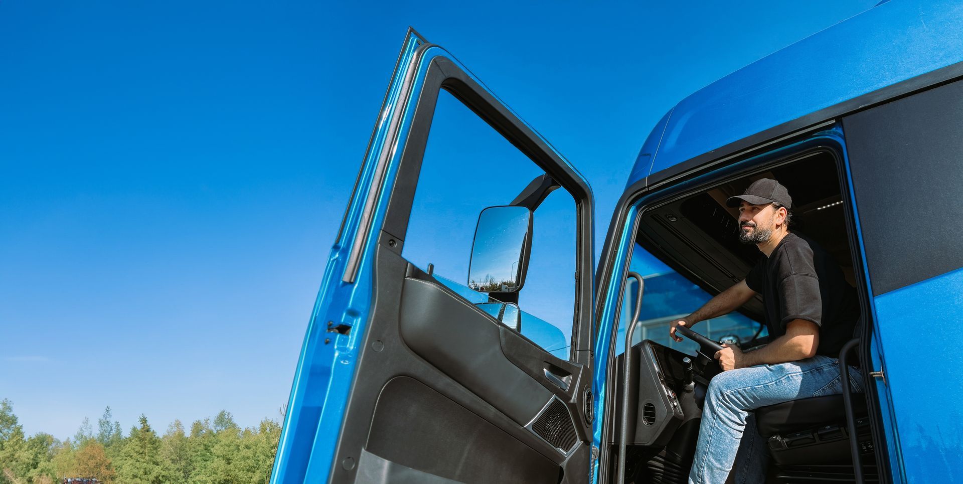 Man driving a blue semi-truck with the door open against a bright blue sky.