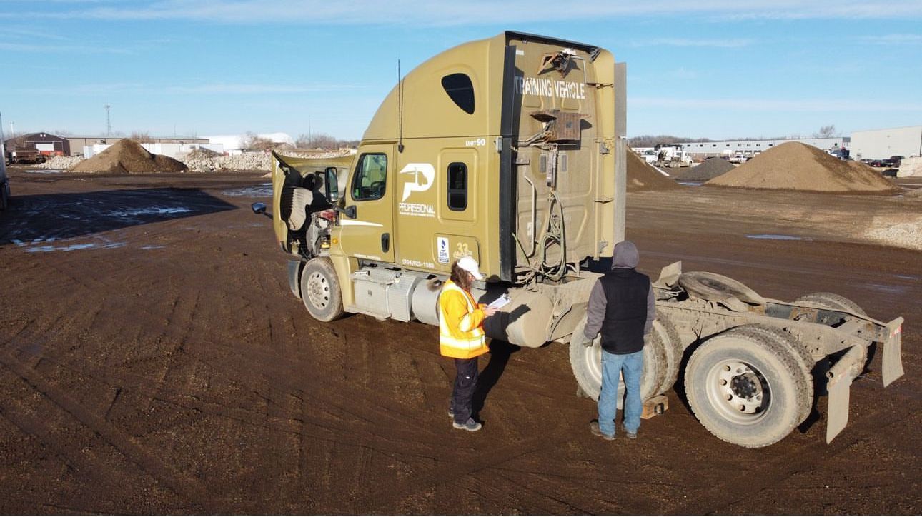 Two people in high-visibility gear inspect a tan semi-truck parked in a muddy, open lot.