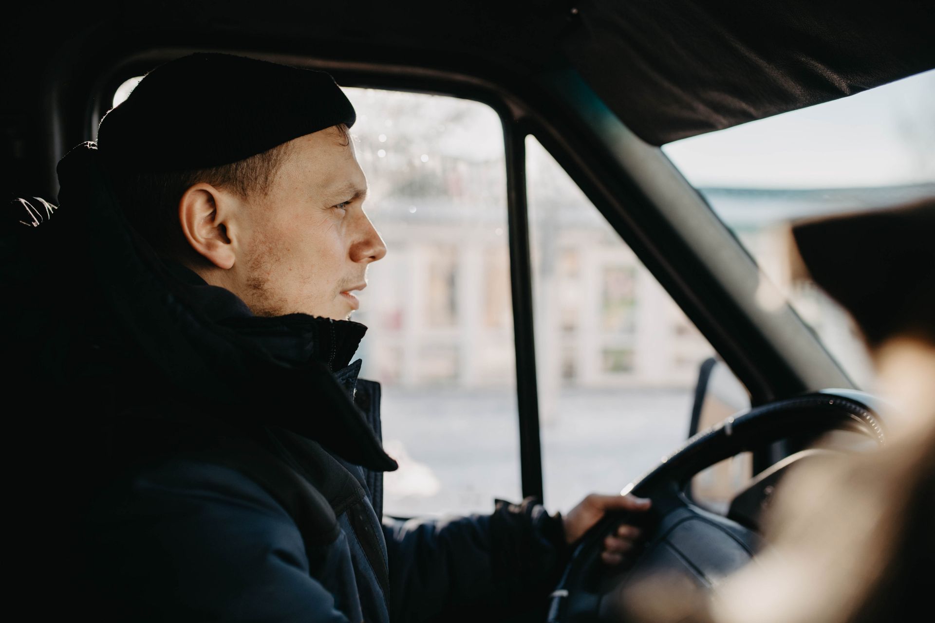 Man driving a vehicle, wearing a black cap and jacket. Sunlight reflects on the window.