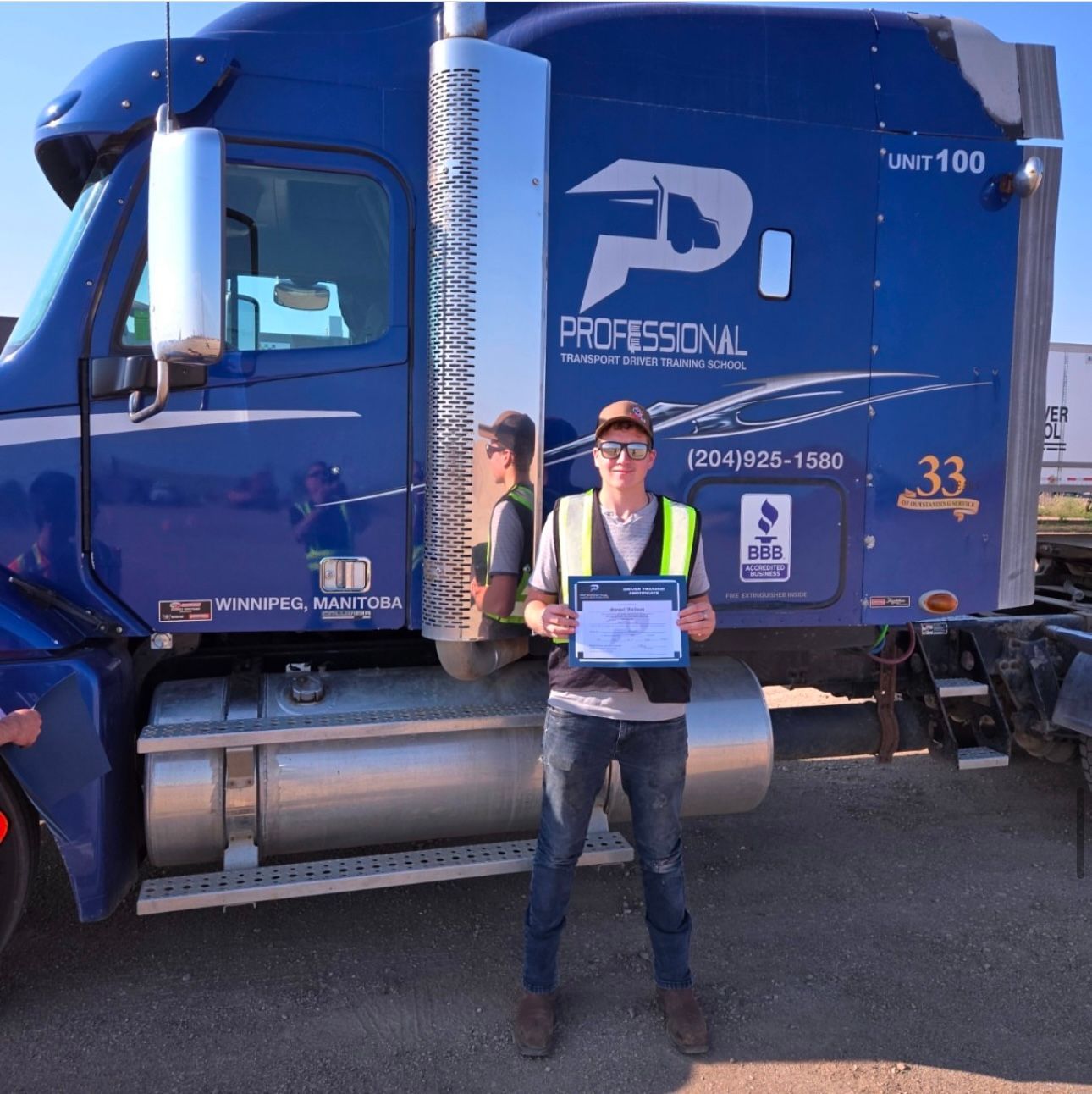 A person wearing a high-visibility vest stands in front of a blue semi-truck, holding a certificate.