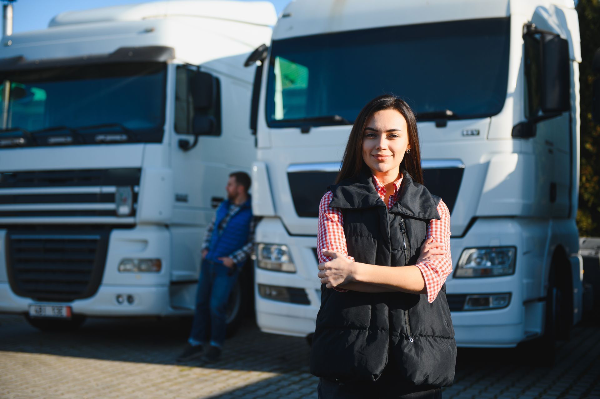 Woman in vest stands in front of two white semi-trucks, arms crossed, smiling. Man in background.