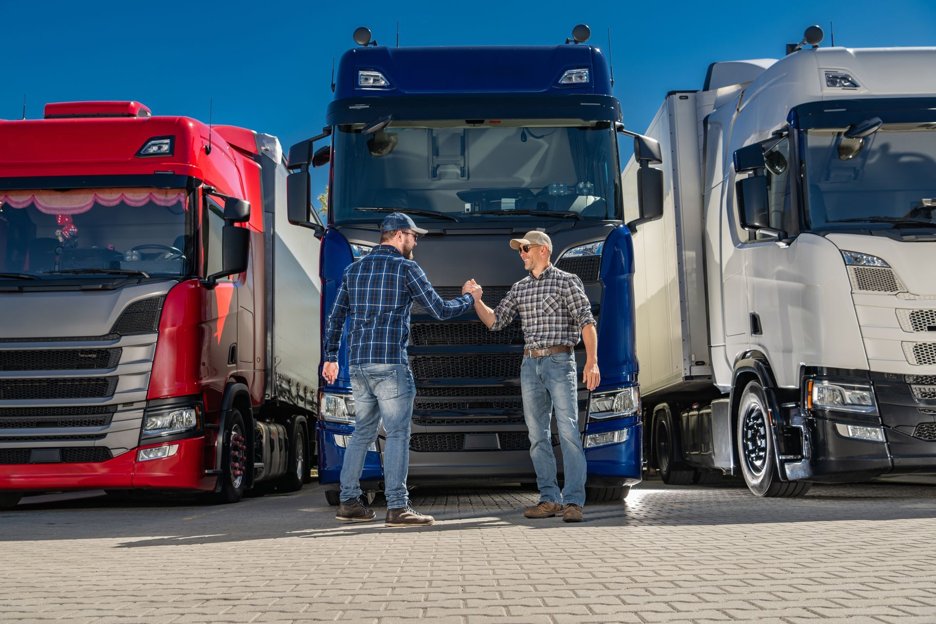 Two men fist-bump in front of three parked semi-trucks, red, blue, and white, on a sunny day.