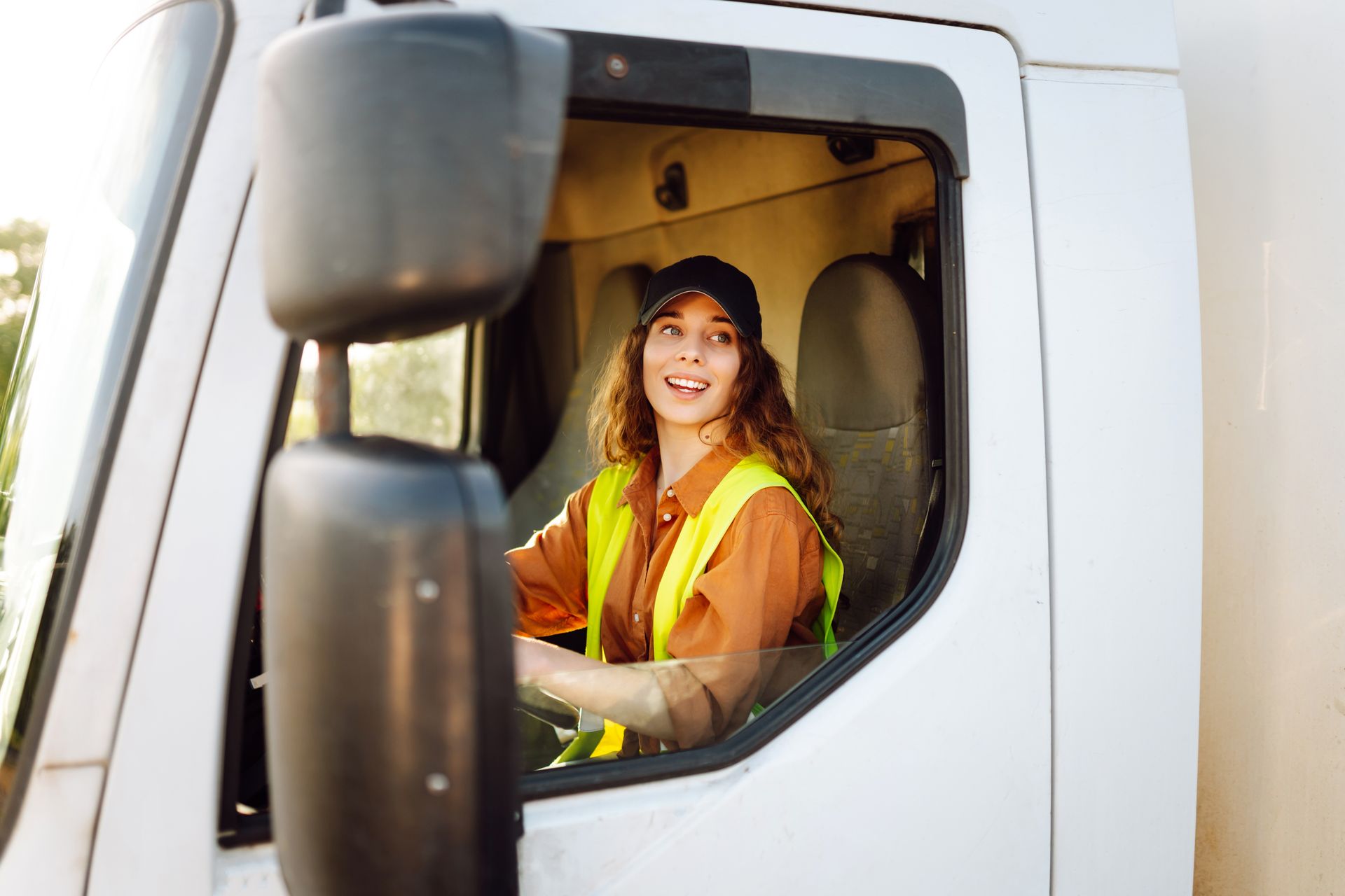 Woman driving a white truck, smiling, wearing a safety vest and cap.