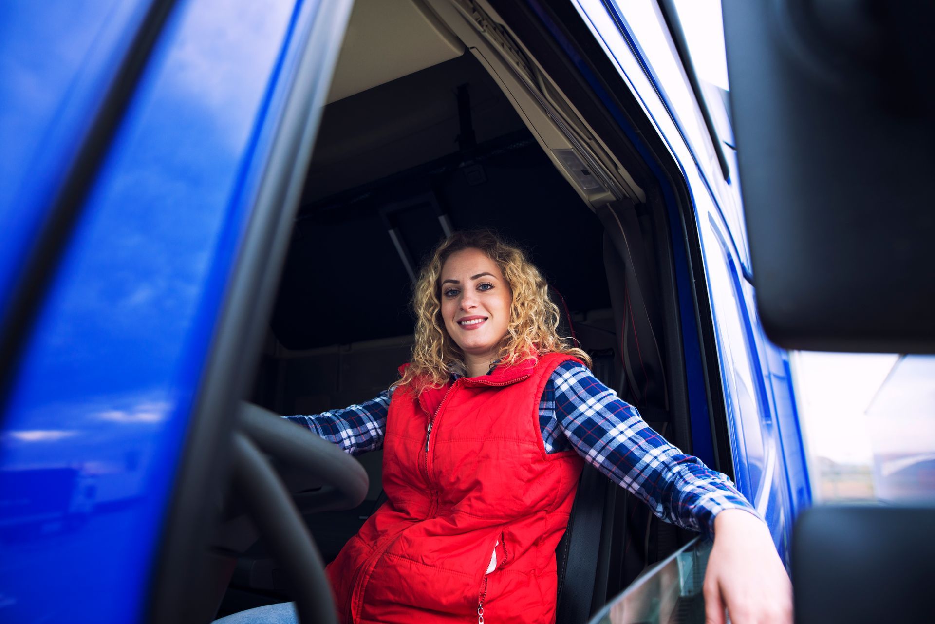 Blonde woman in red vest smiles from blue truck cab, holding the steering wheel.