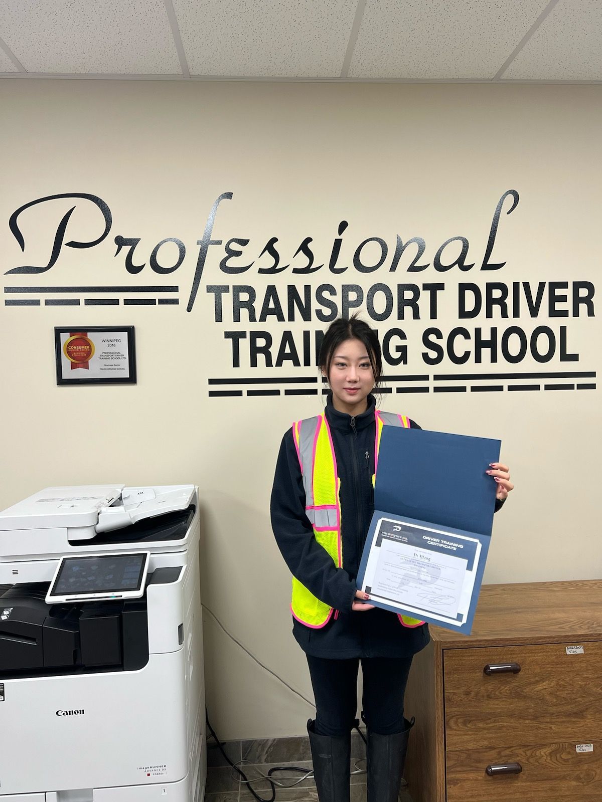 A student in a high-visibility vest holds a graduation certificate in front of a Professional Transport Driver school sign.