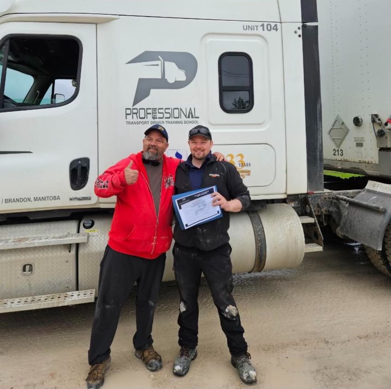 Two people standing in front of a white semi-truck, one giving a thumbs up and the other holding a certificate.