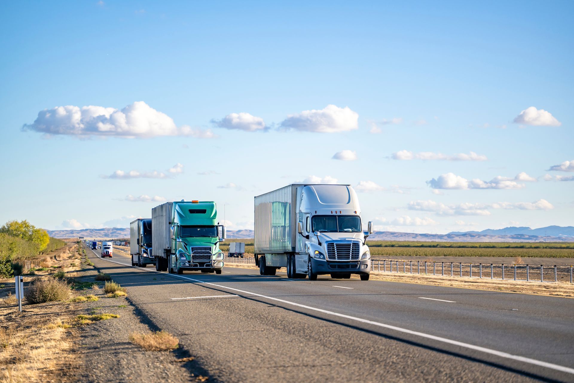 Semi-trucks driving on a highway, under a partly cloudy sky.