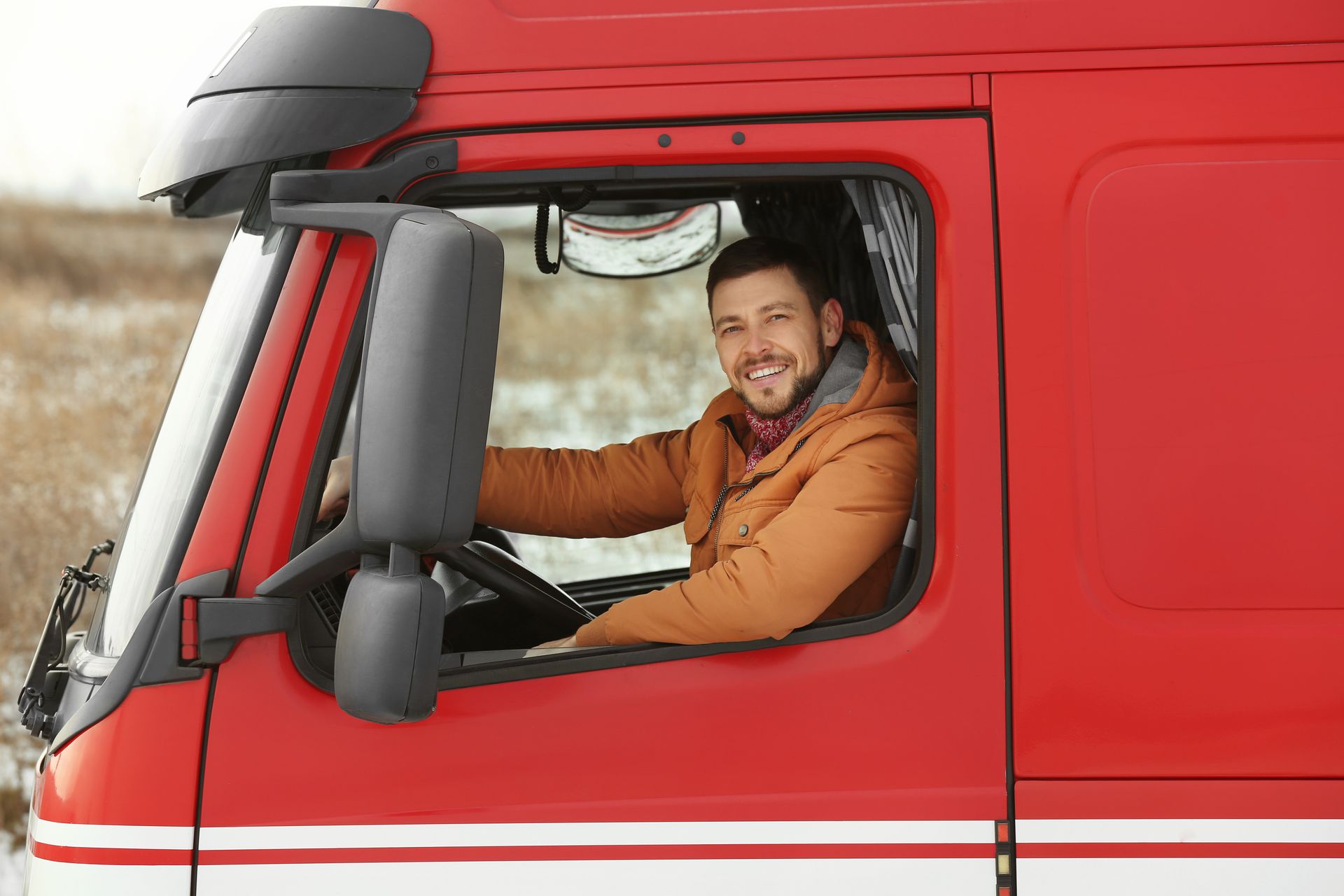 Man smiling in the cab of a red semi-truck, outdoors.