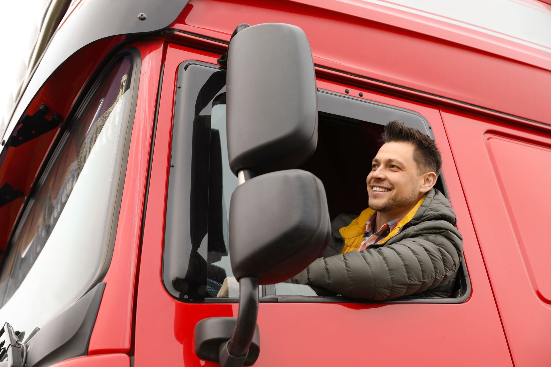 Smiling truck driver in a red semi-truck, looking out the open window.