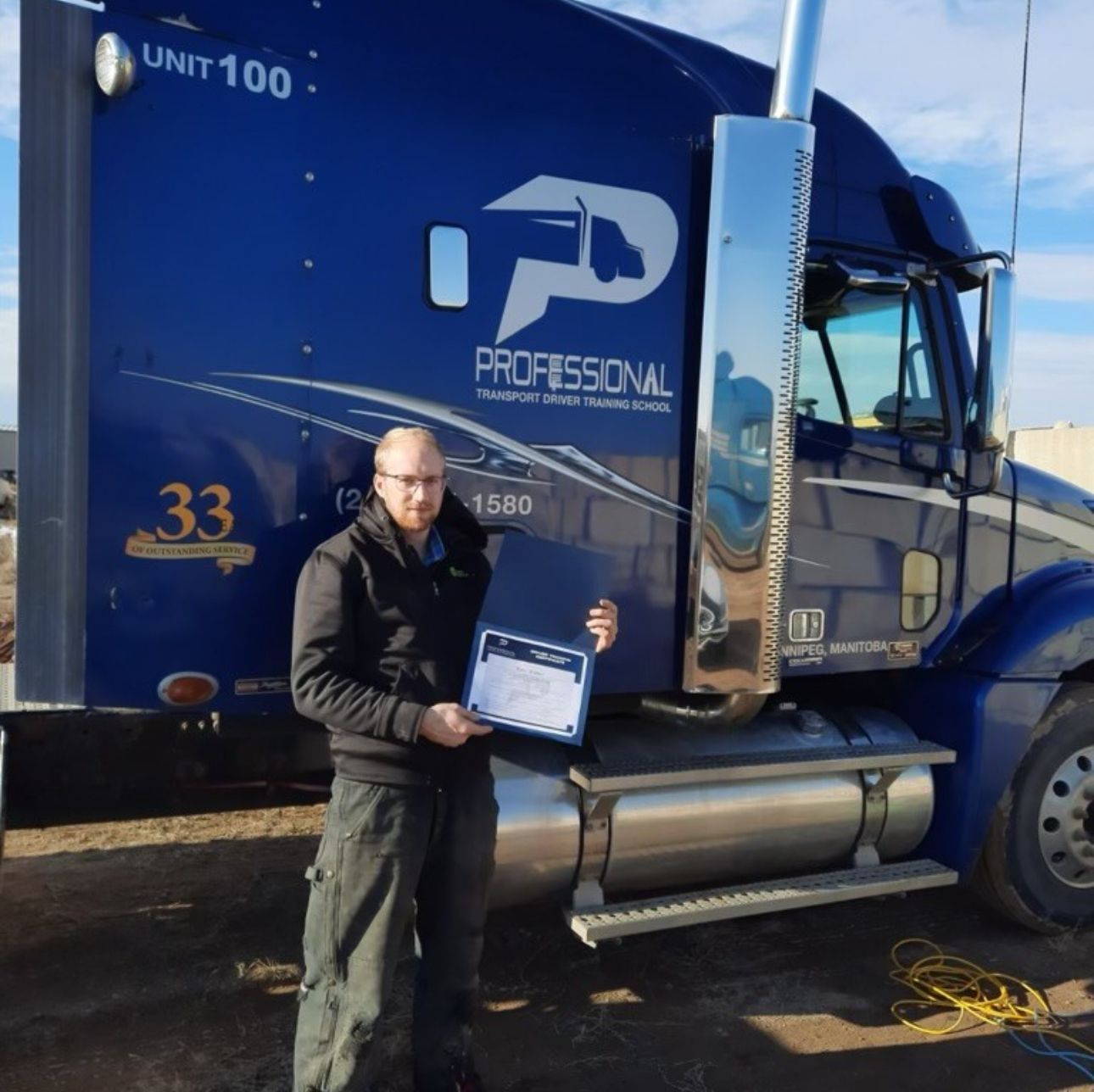 A person stands in front of a blue semi-truck, holding a certificate.