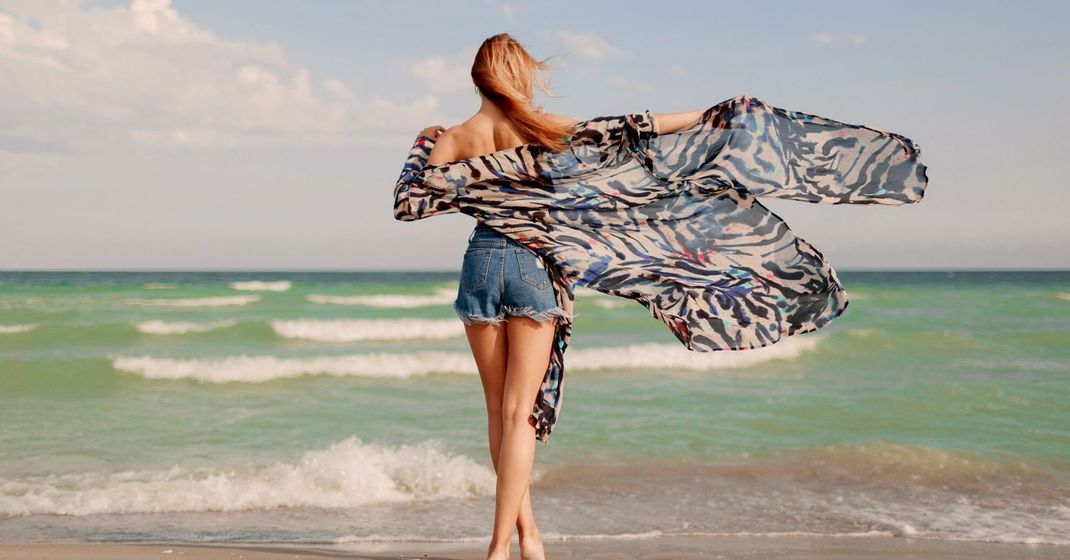 A woman is standing on a beach holding a scarf around her neck.