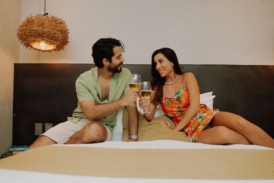 A man and a woman are sitting on a bed toasting with wine glasses.