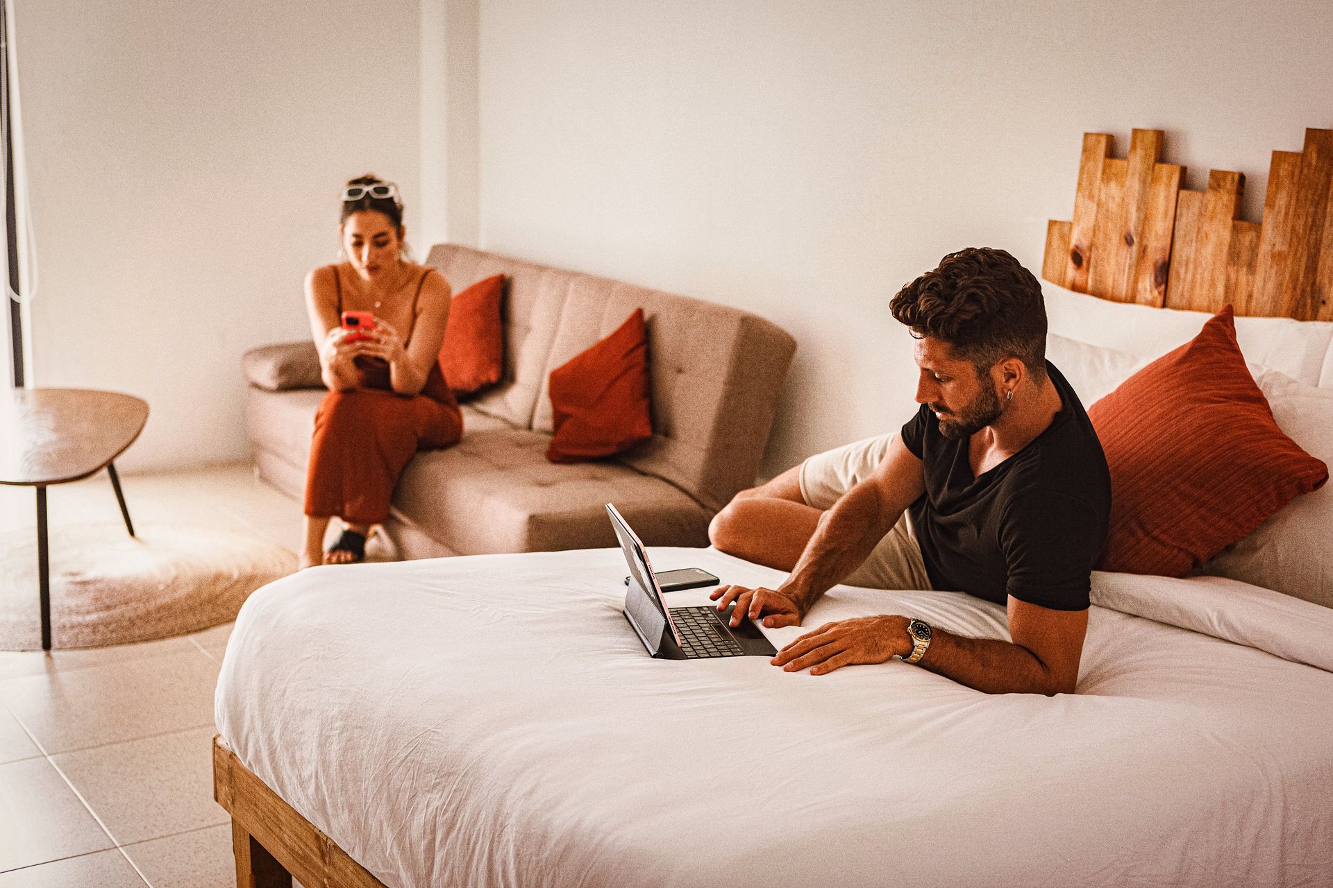 A man is sitting on a bed using a laptop while a woman sits on a couch.