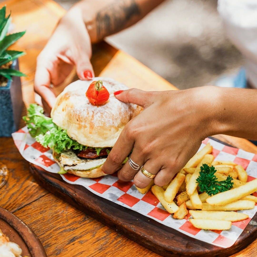 A person is holding a hamburger and french fries on a wooden cutting board.