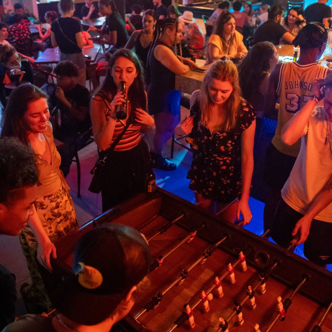 People playing foosball at a crowded bar; others are drinking, smiling, illuminated with blue and orange lights.