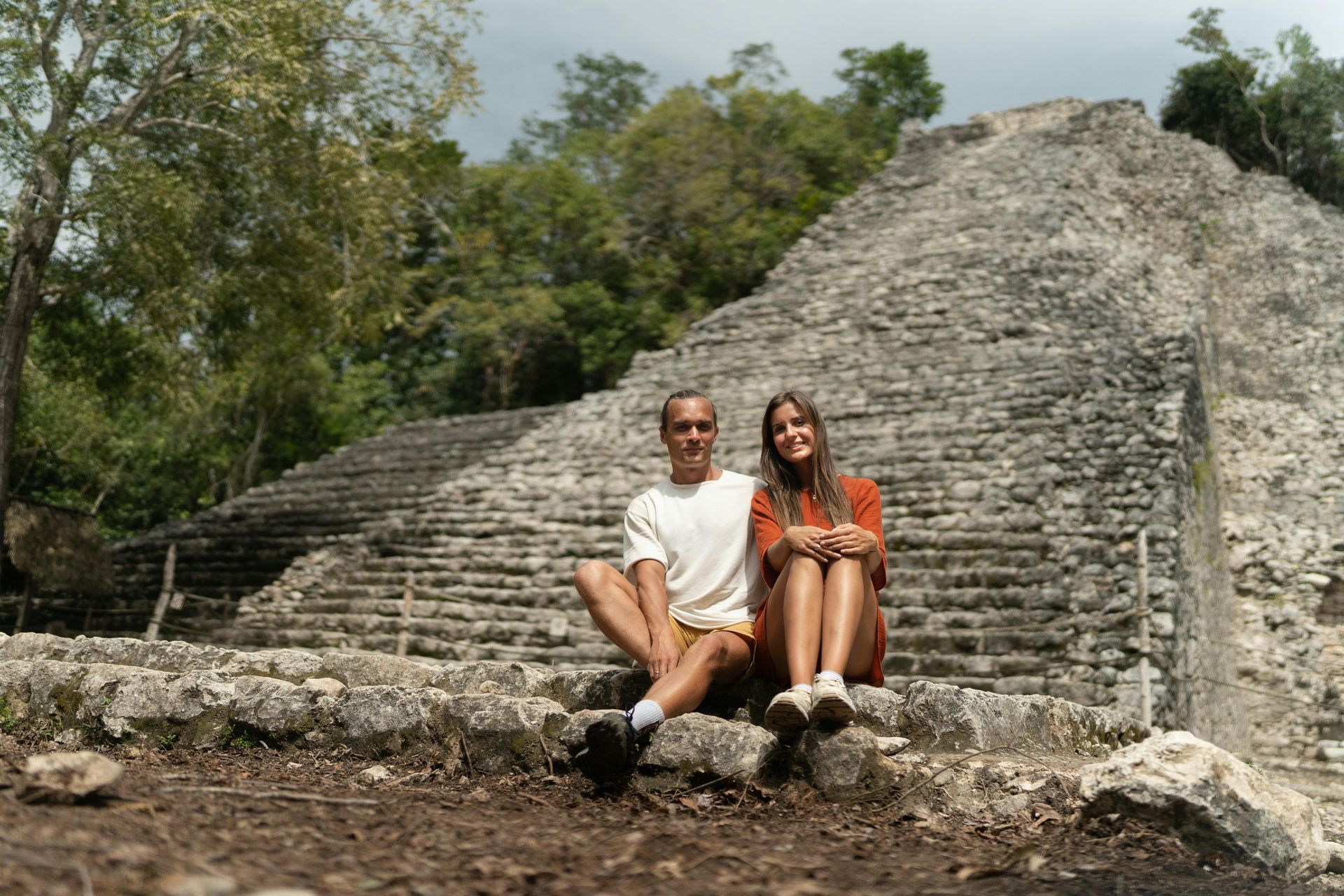Couple sits on steps of ancient Mayan pyramid, green trees in background.