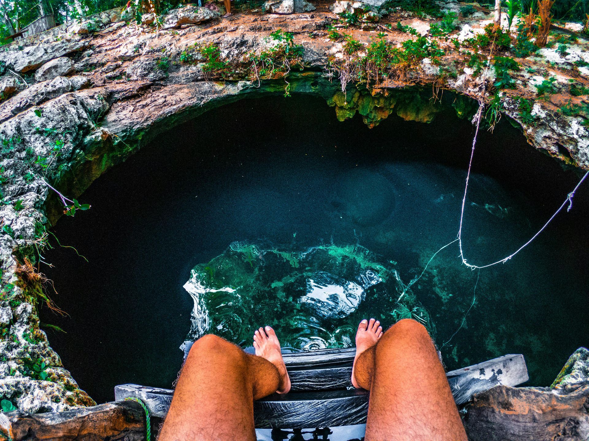 Person's legs dangling over a cenote. Blue-green water in a limestone sinkhole.