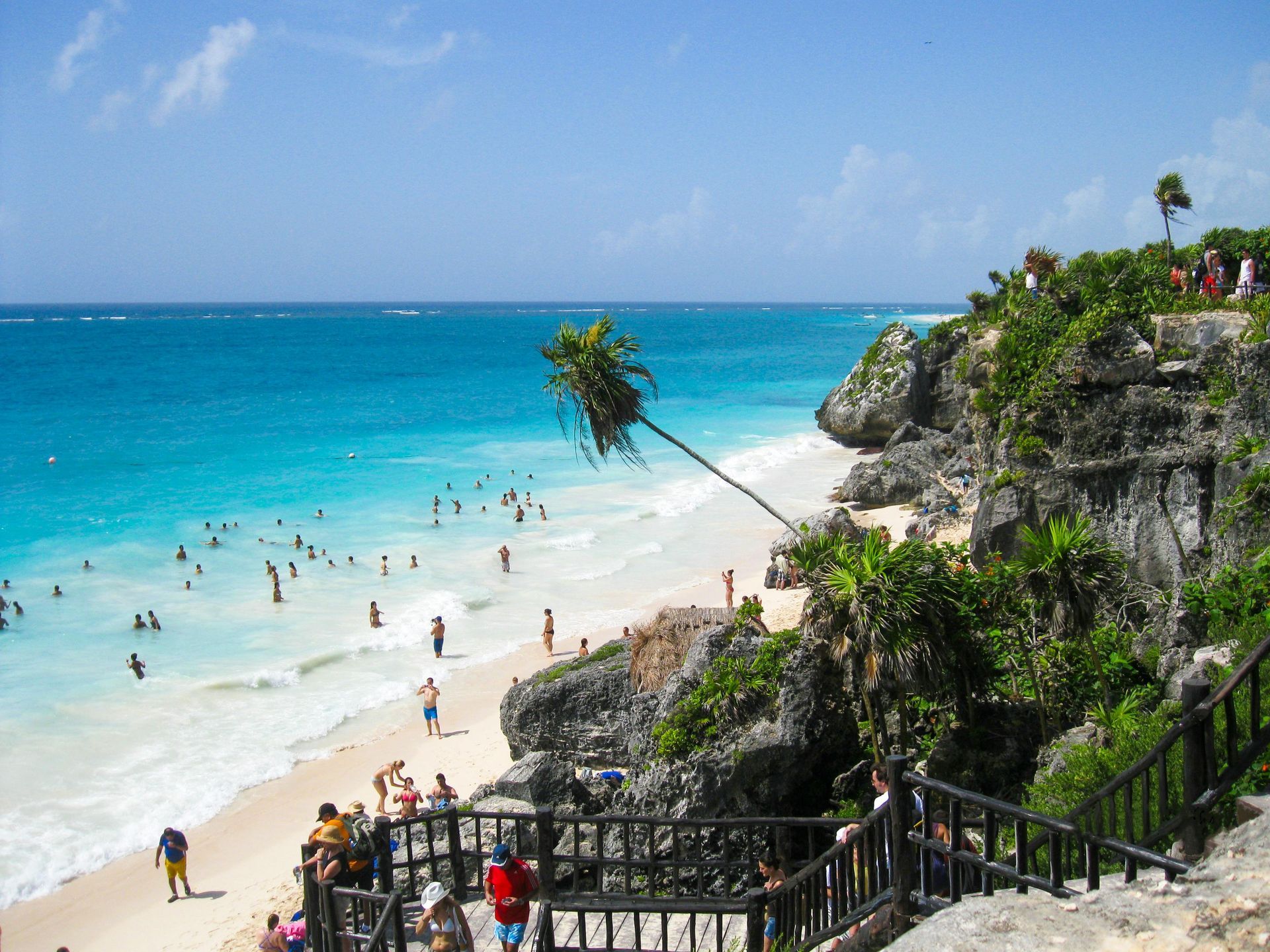 Beach with turquoise water, white sand, and people swimming. Cliffside with vegetation. Sunny sky.