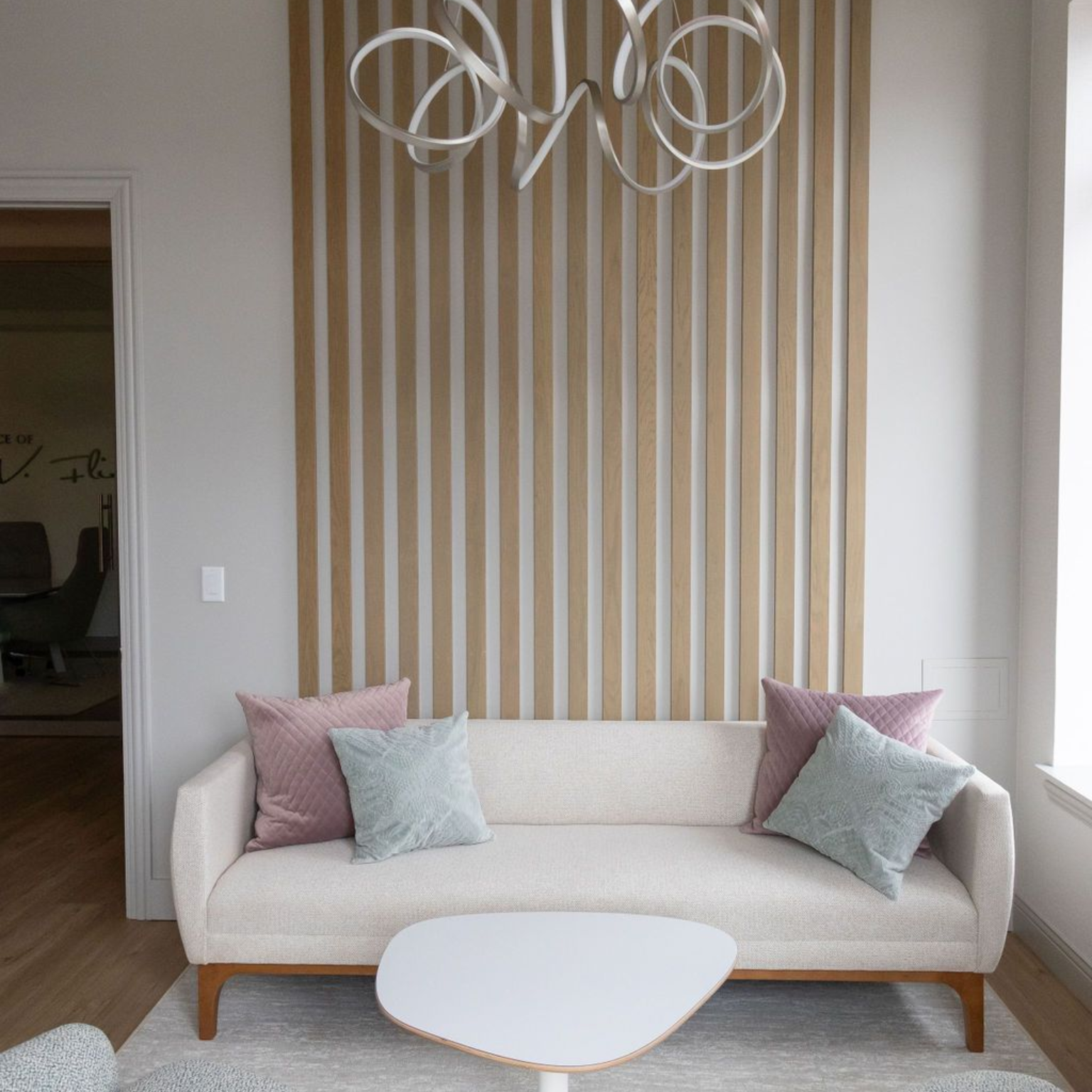 Beige sofa with pink and teal pillows, in front of a vertical wood panel, under a modern light fixture.