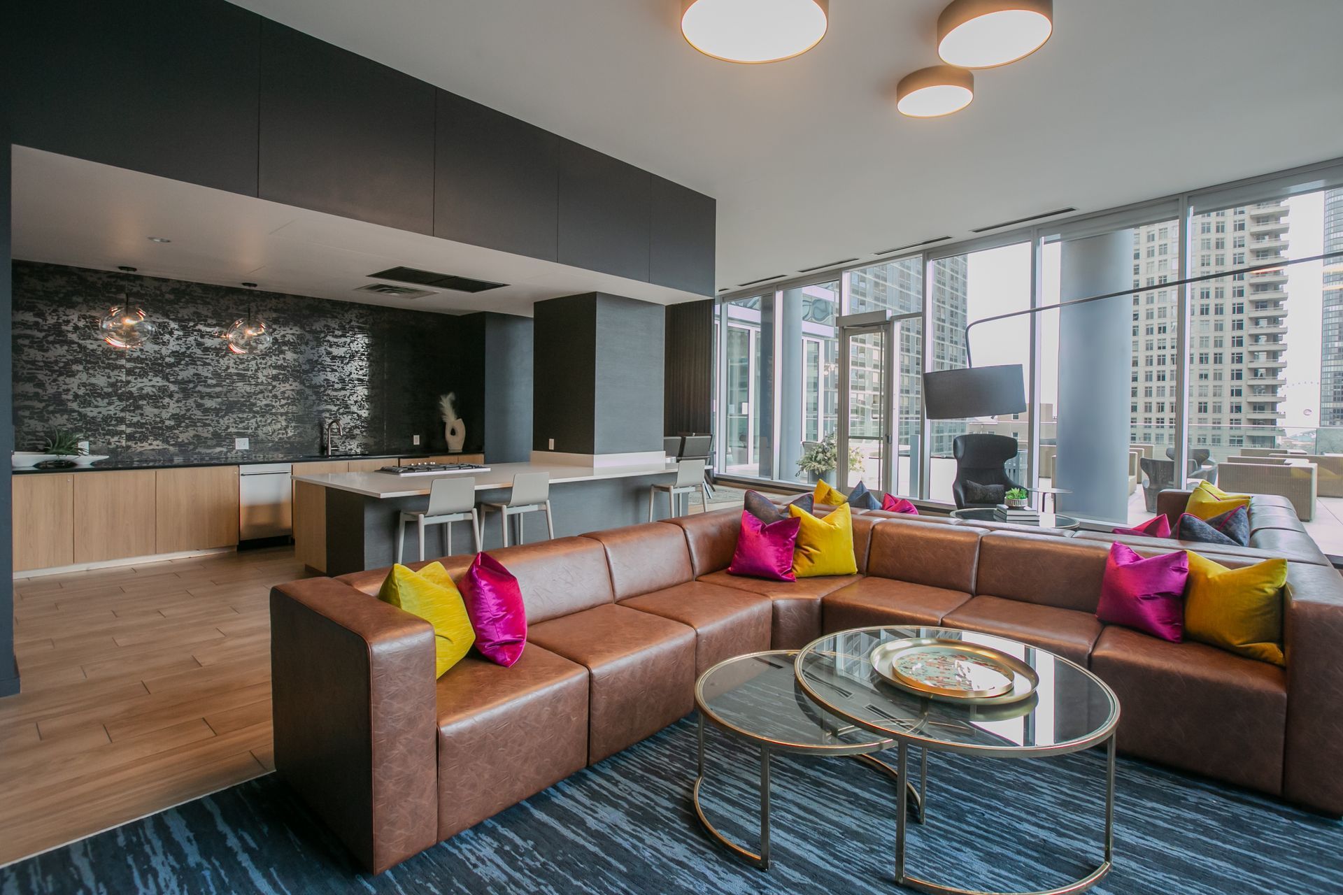 Lounge with brown leather sectional, colorful pillows, kitchen area, and city view.