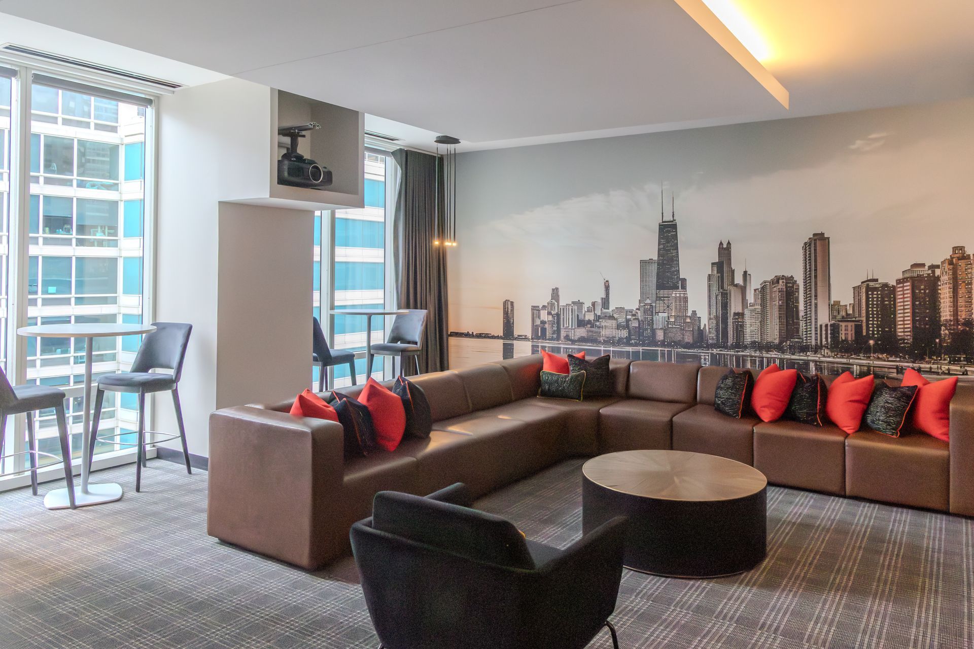 Lounge area with brown sectional, Chicago skyline mural, round coffee table, bar stools, and city view.