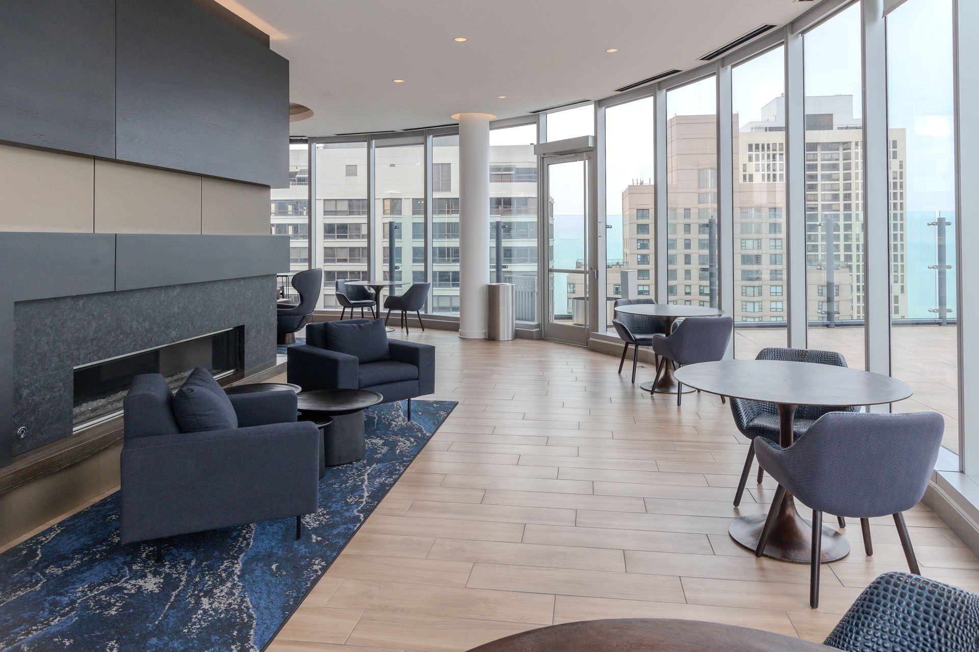 Modern lounge with city view, featuring armchairs, tables, and a fireplace. Light wood floors and large windows.