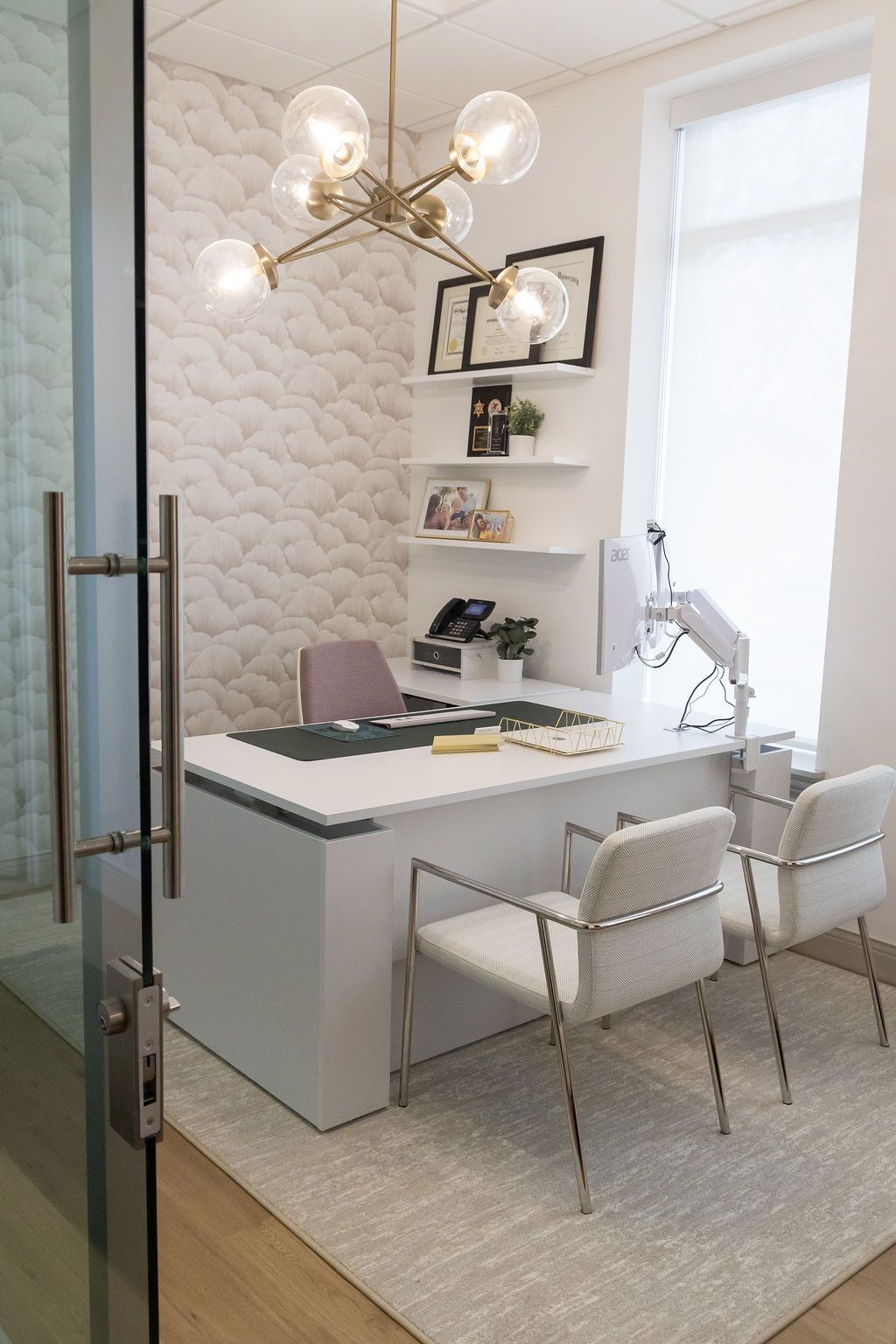 Office with white desk, chairs, and patterned wallpaper. Gold chandelier hangs above.