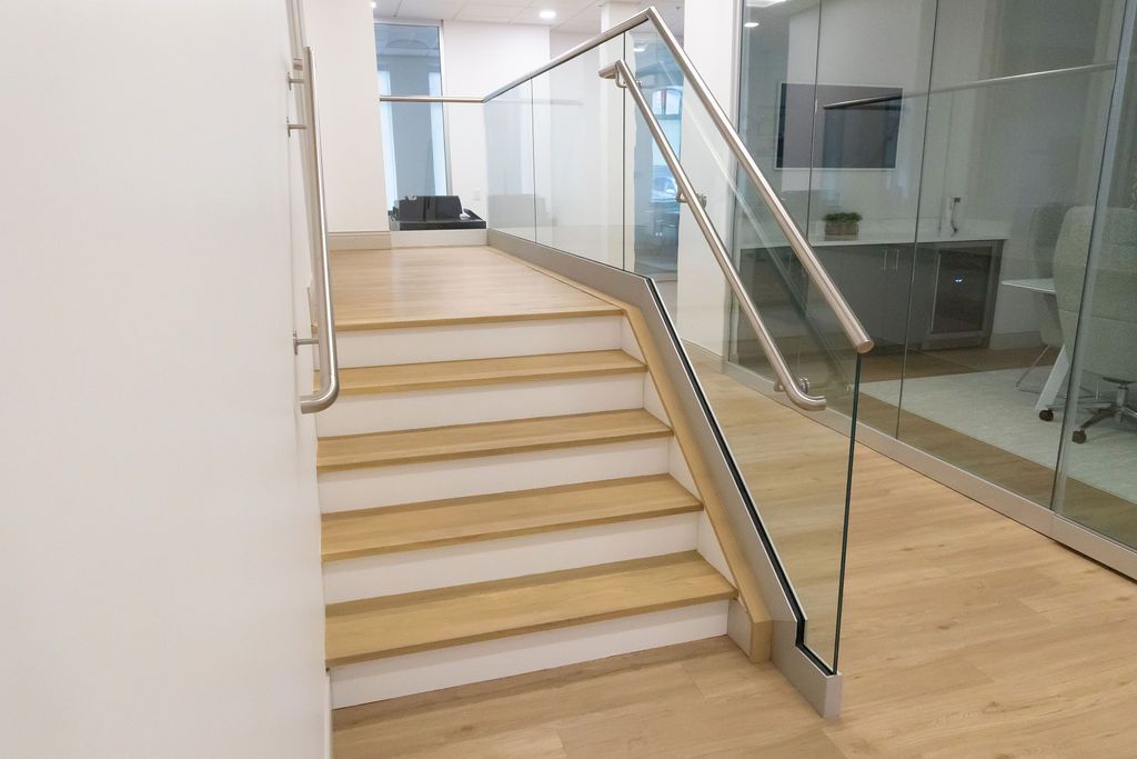 Staircase with wooden steps and glass railing, leading to a hallway with a desk and white walls.
