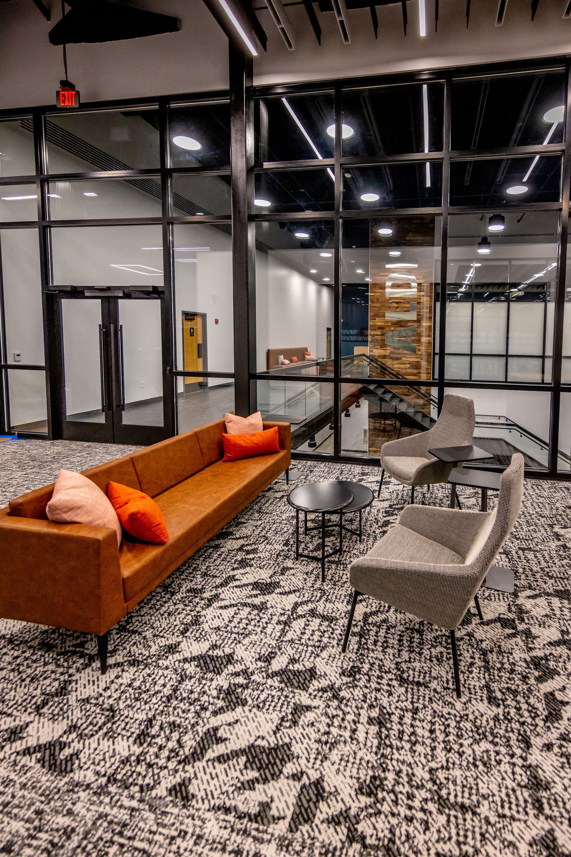 Lounge area with brown sofa, patterned chairs, and black tables in front of a glass wall.