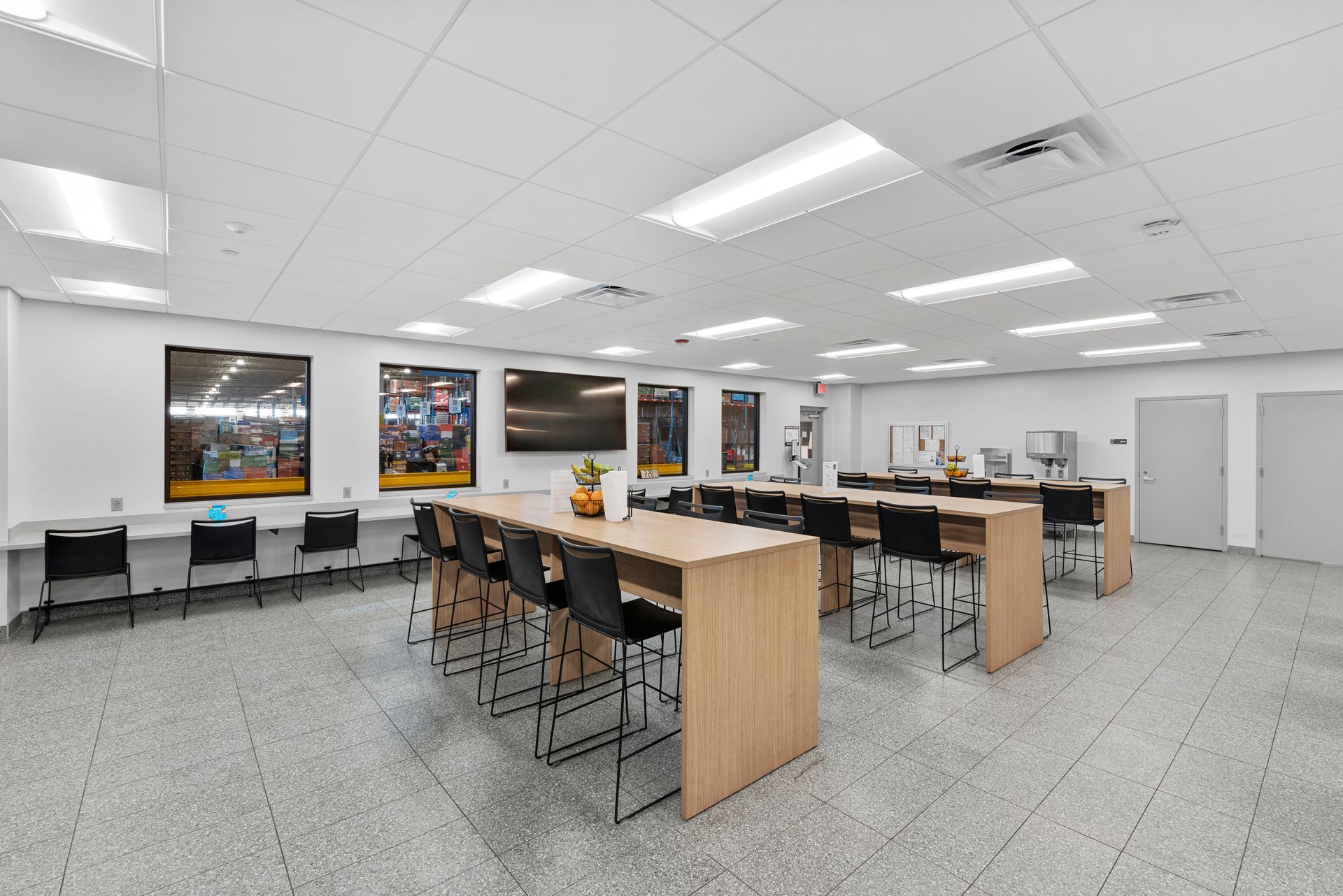 A bright, open room with long tables, chairs, posters, and TVs. Fluorescent lights illuminate the space.