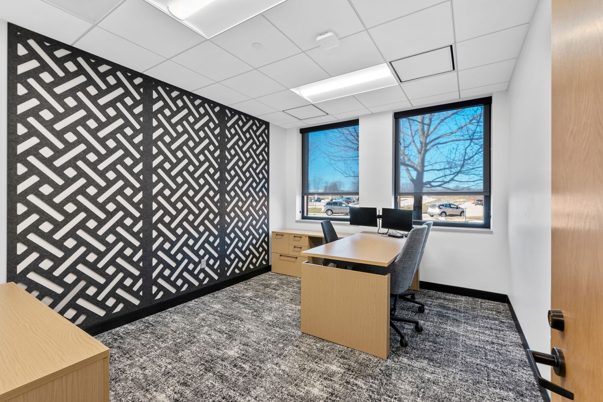 Modern office space with patterned accent wall, two desks, and windows overlooking a parking lot.