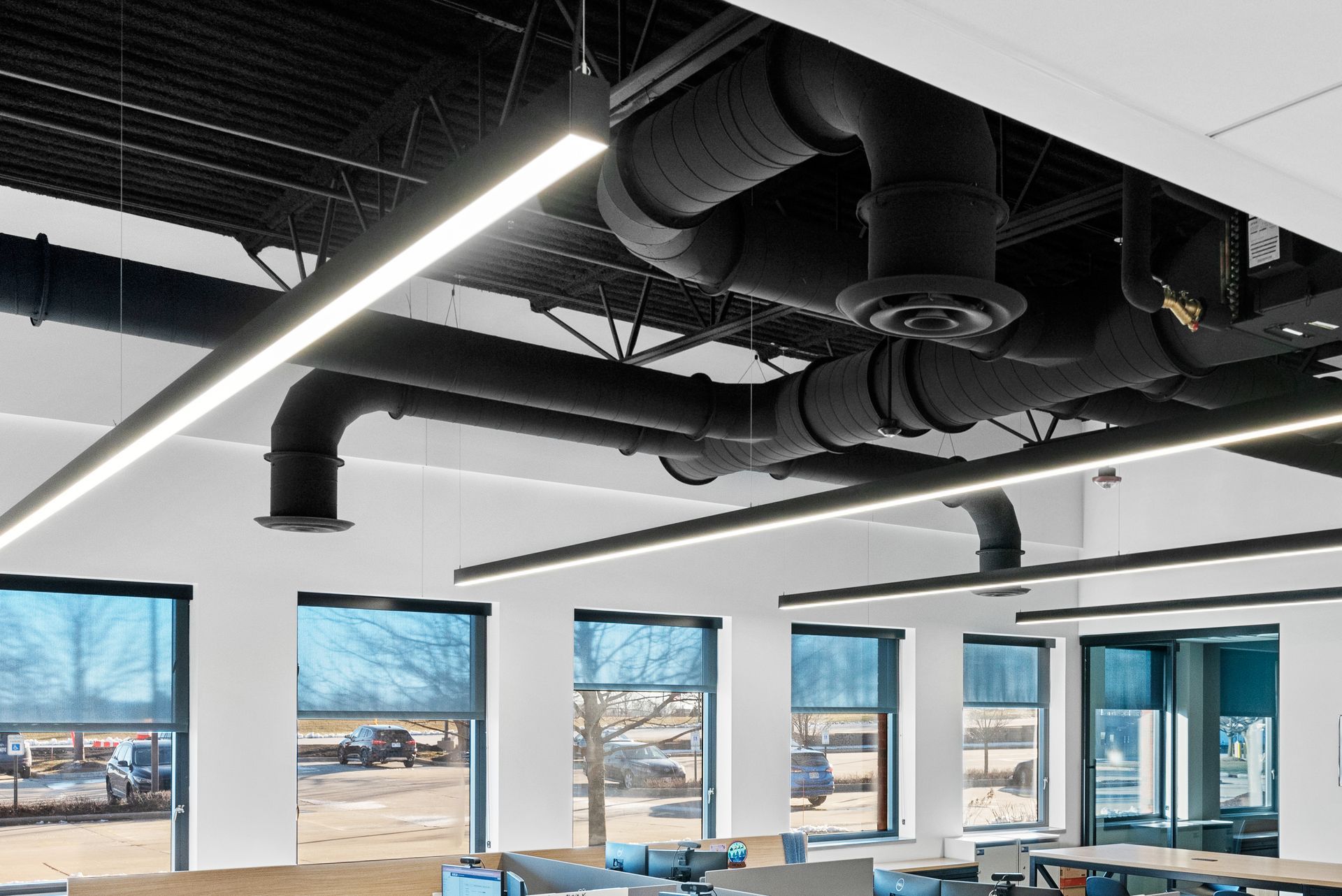 Office interior with black ceiling, linear lights, and exposed ducts. Large windows offer a view of an outdoor scene.