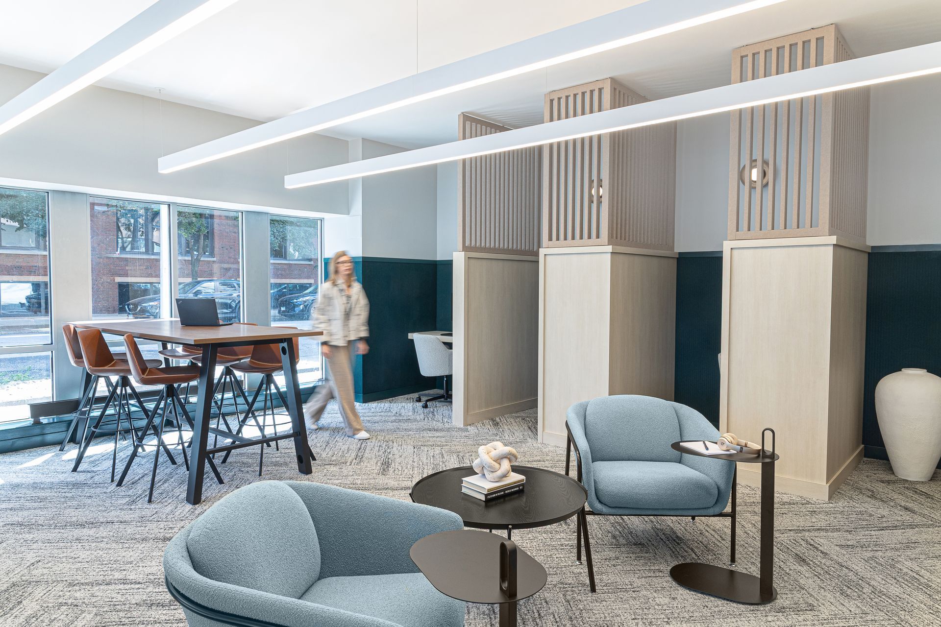 Cozy co-working space: woman walks past seating area with blue armchairs, round table, and high table with laptop.