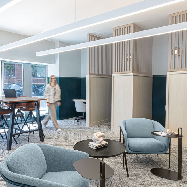 Modern office lounge with blue armchairs, round tables, and a person walking past a high-top table.