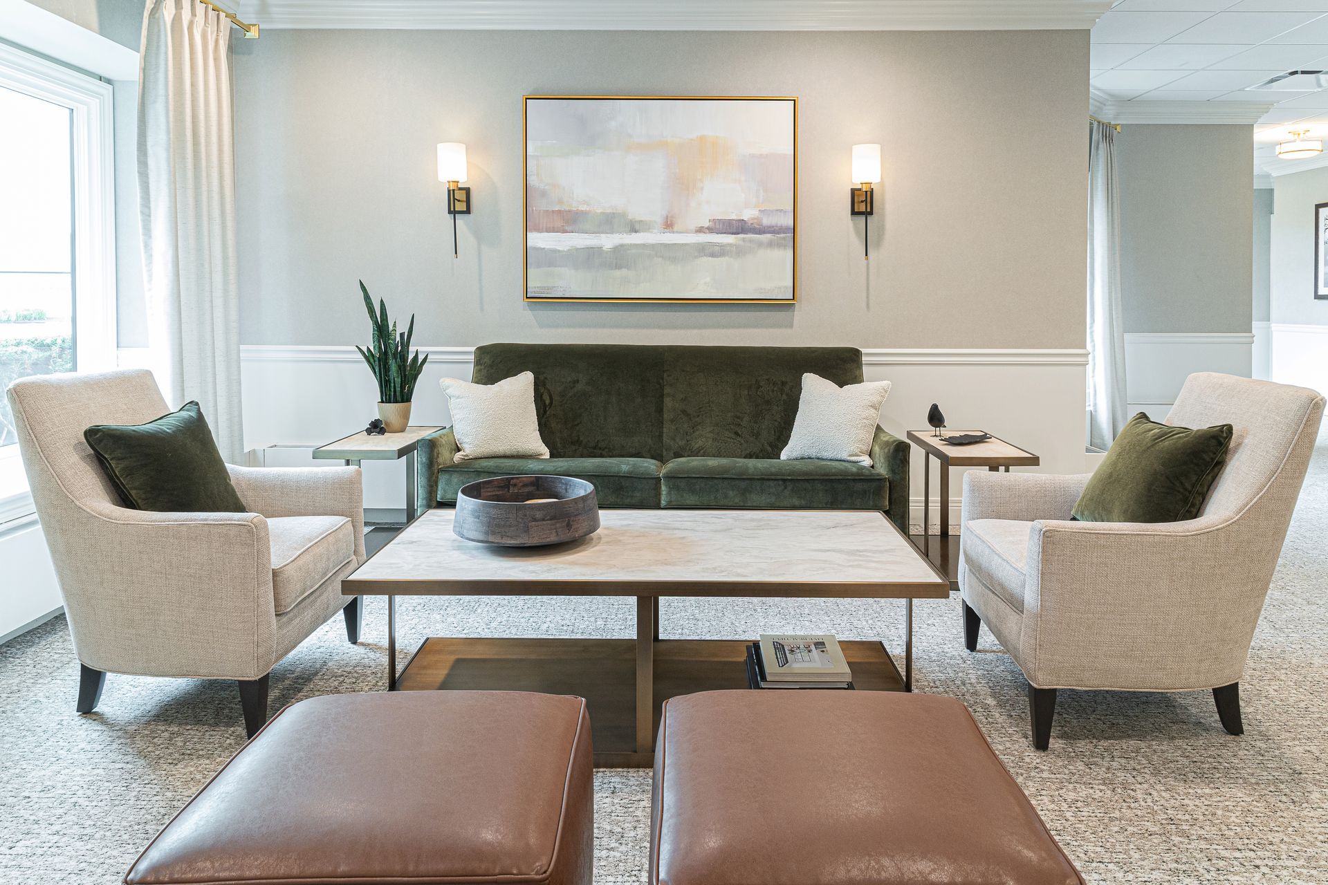 Lounge area with green sofa, cream armchairs, coffee table, and tan ottomans.  Artwork and sconces on light gray wall.