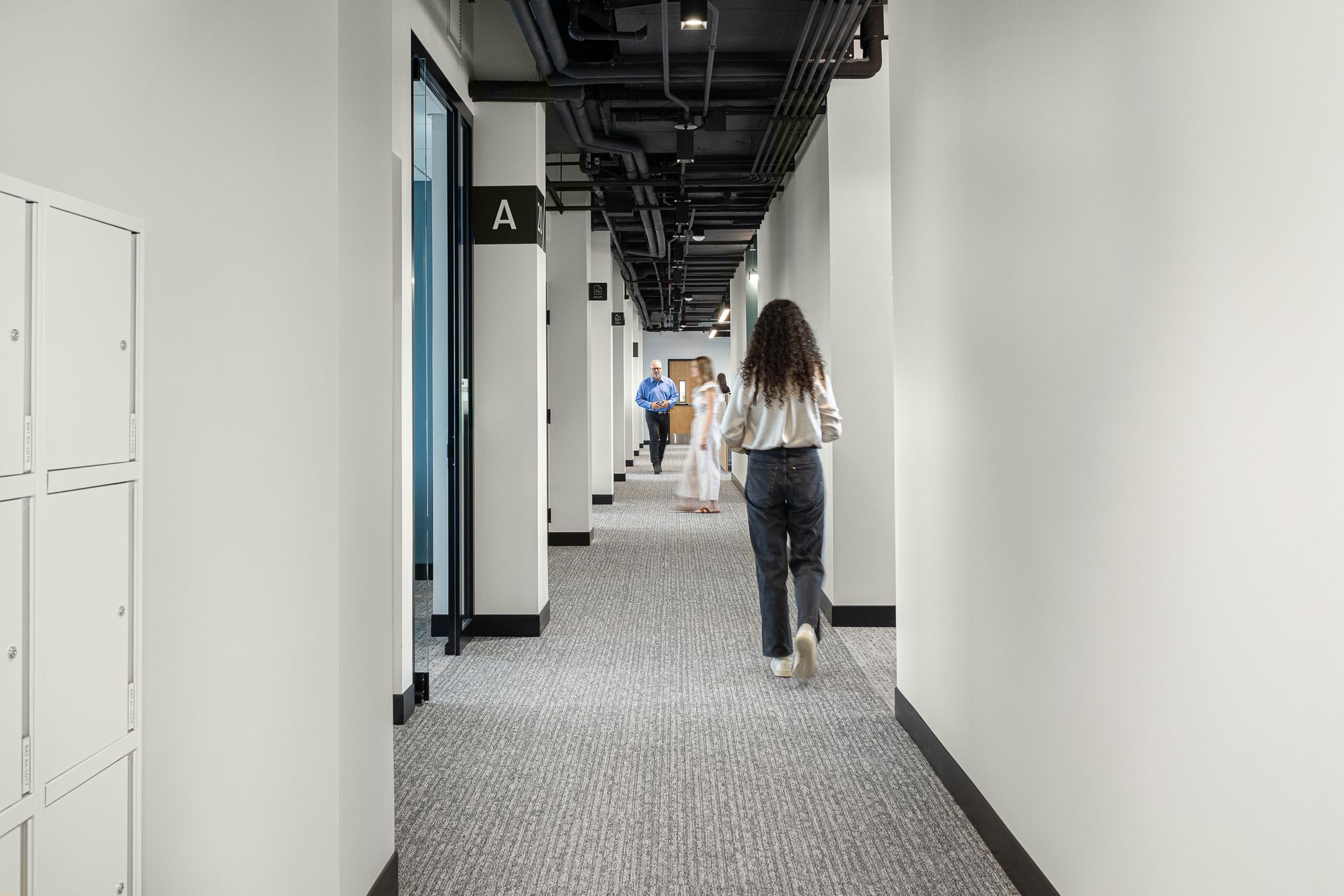 Woman walking down a hallway with white walls and a carpeted floor. Other people are further down the hall.