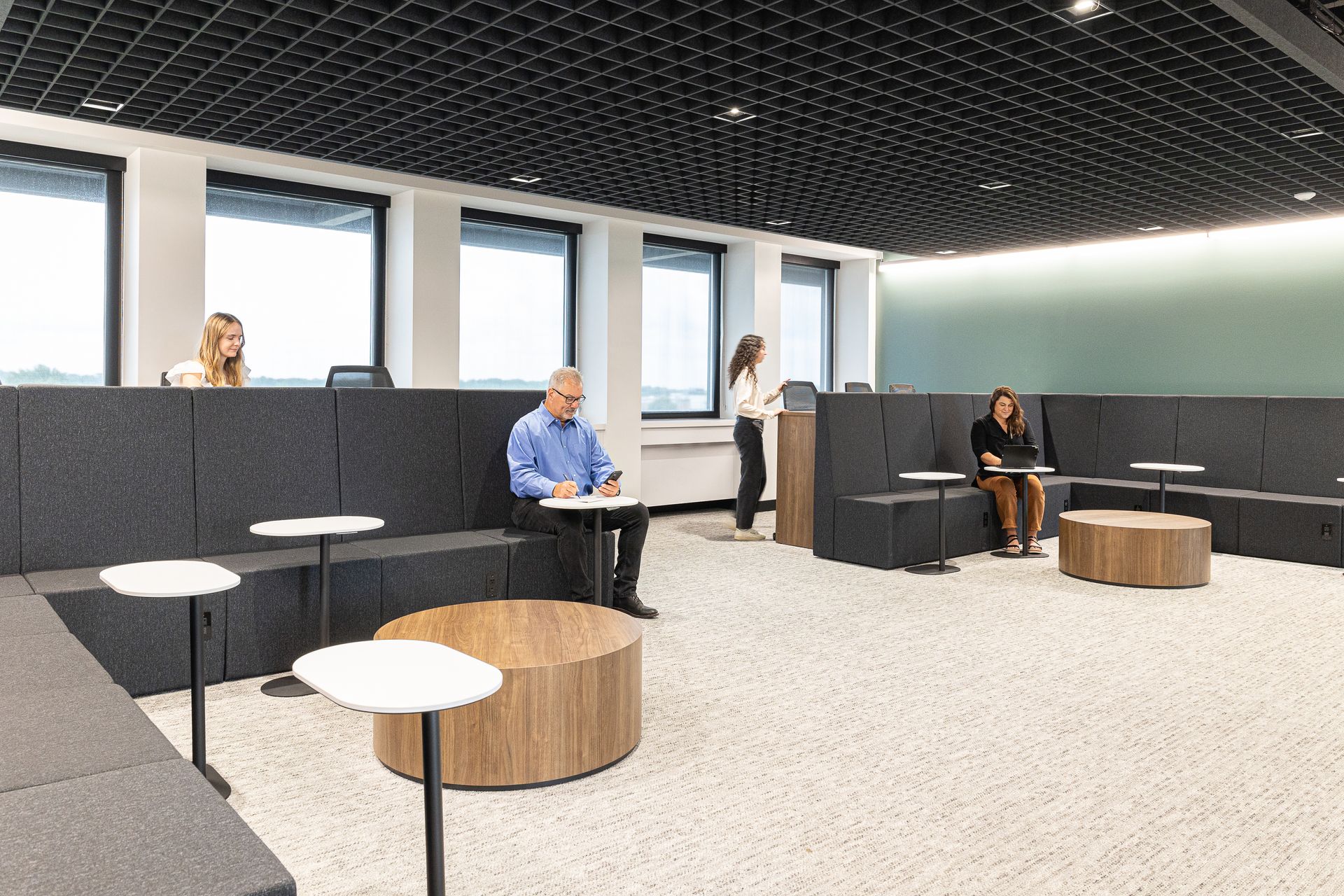 People in an office lounge with modular seating, small tables, and a textured ceiling.