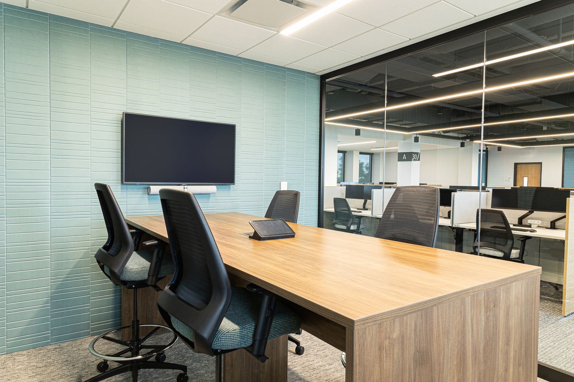 Conference room with wooden table, chairs, and large TV on a blue textured wall. Interior office space visible through glass.