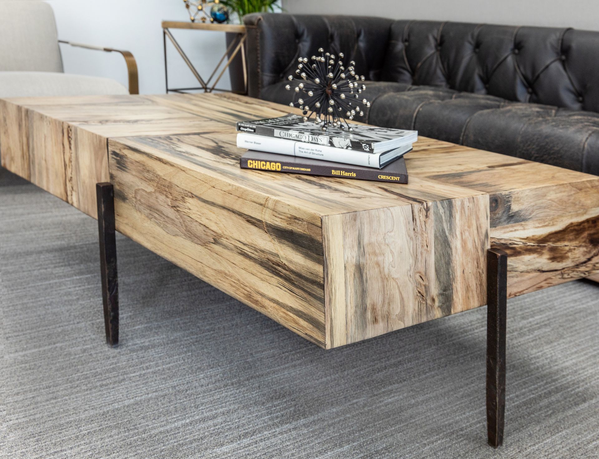 Wooden coffee table with dark metal legs, books, and decorative sculpture. Black leather couch in background.